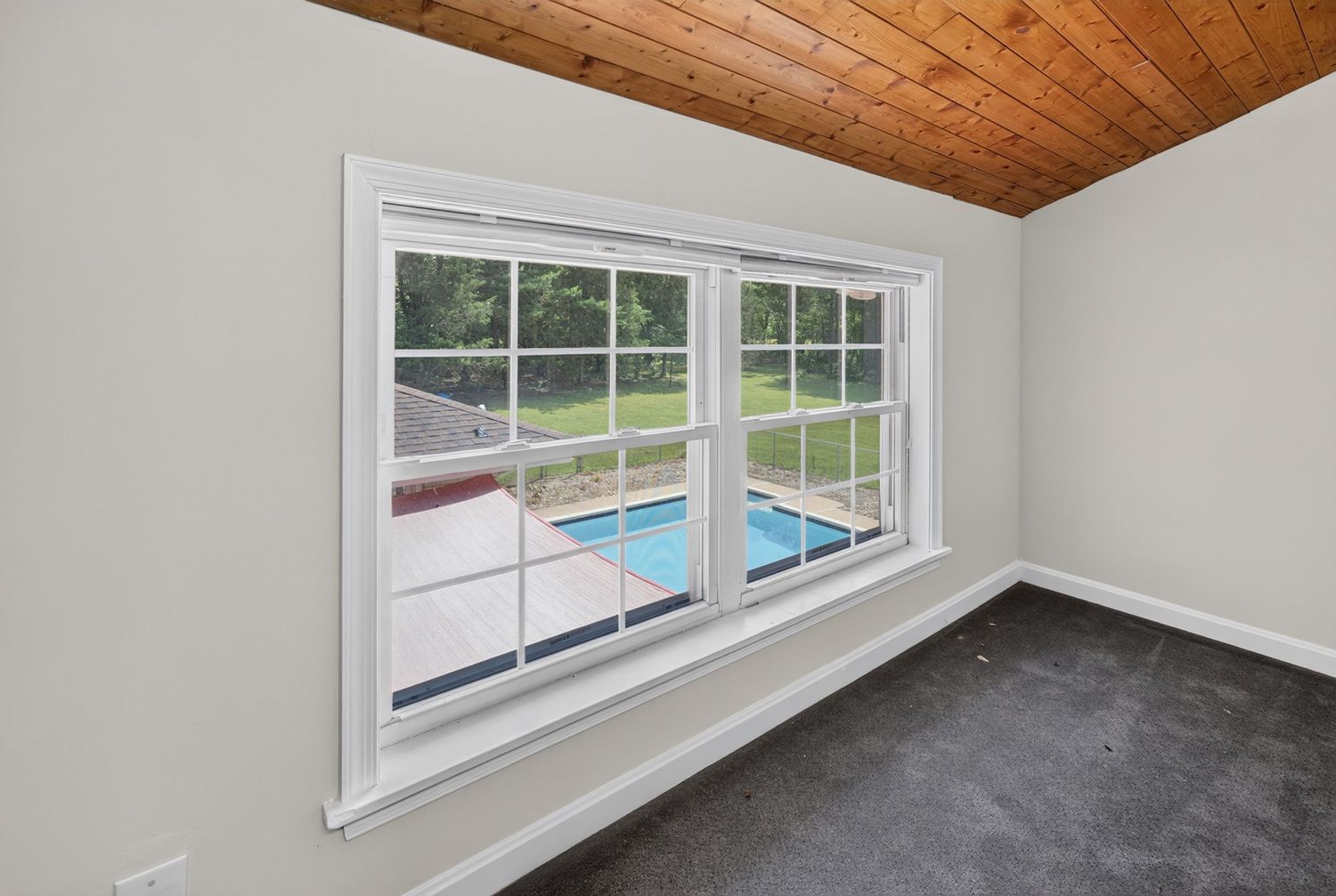 Empty room with white trim, wood ceiling, and large window overlooking a backyard pool and green lawn.