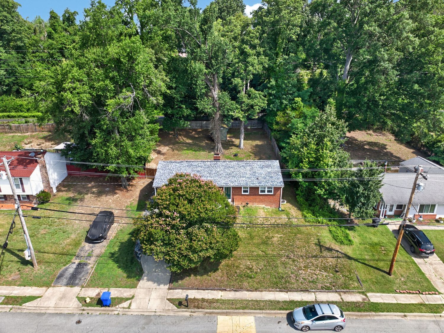 Aerial view of a brick ranch home surrounded by mature trees on a corner lot with green lawn.