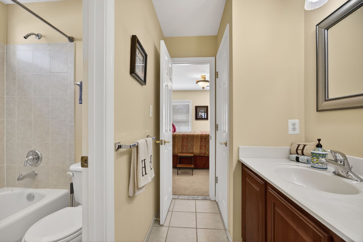 Bright bathroom with tiled tub surround, wooden vanity, white countertop, and doorway leading to an adjacent bedroom.