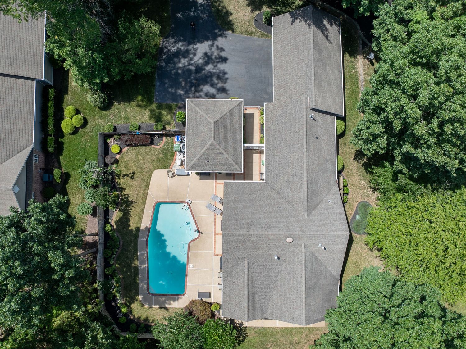 Aerial view of a suburban home with a backyard swimming pool, surrounded by lush green trees and a gray shingled roof.