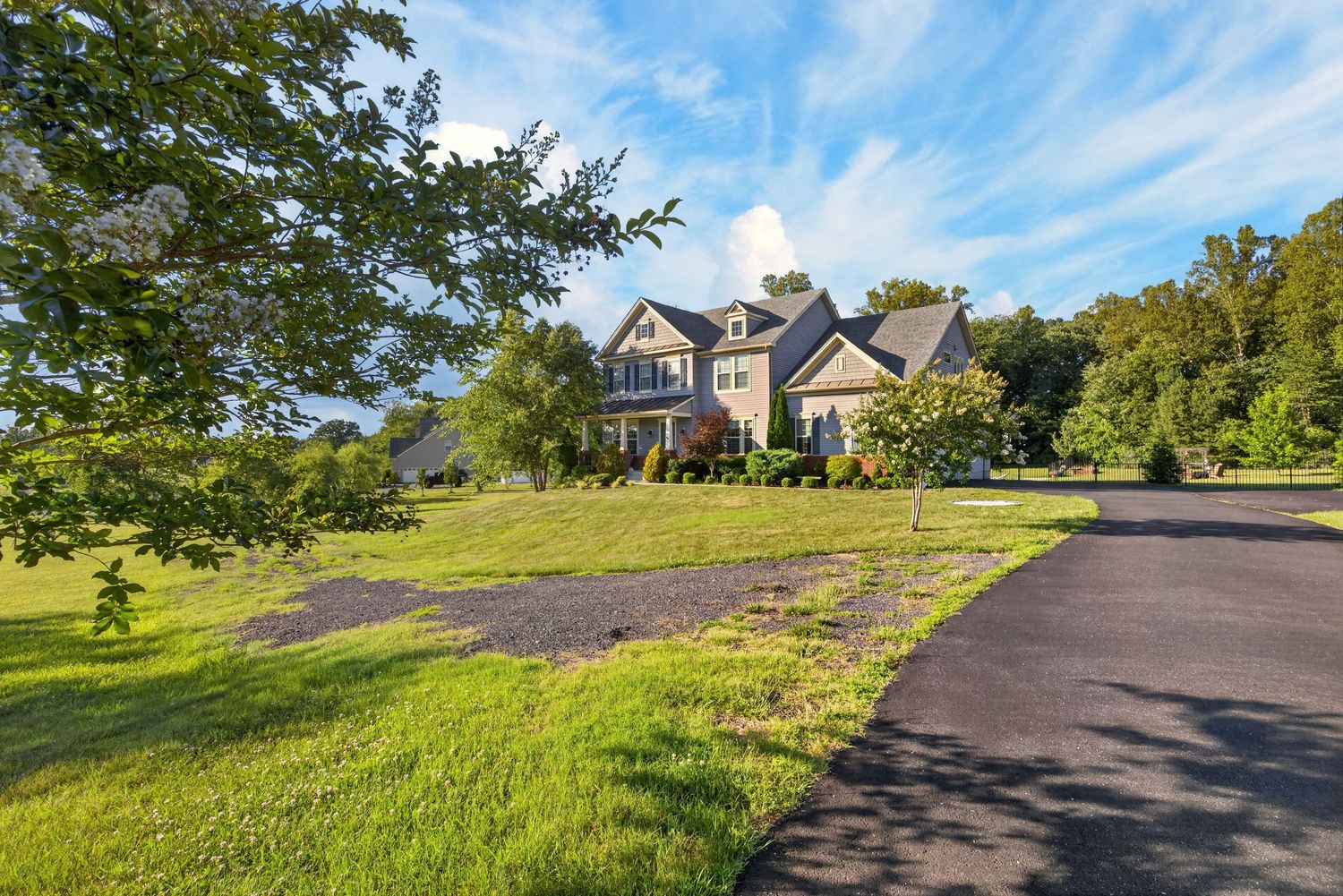 Large suburban home with manicured lawn, paved driveway, and lush trees under a bright blue sky.