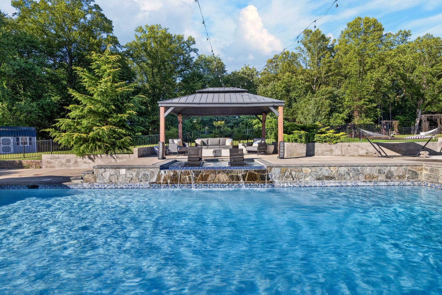 Luxury outdoor pool with waterfall feature, stone patio, and elegant gazebo surrounded by lush green trees.
