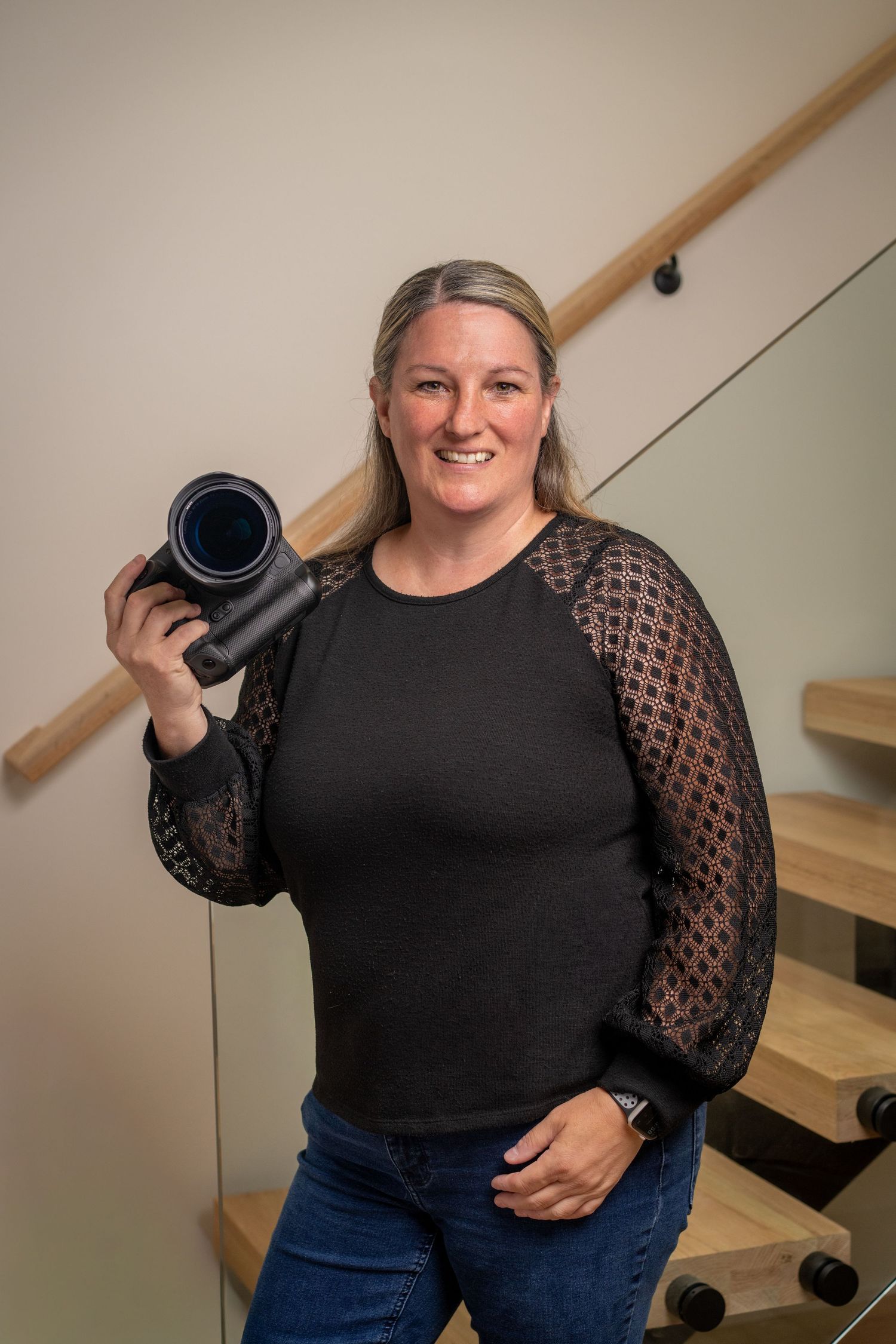 Smiling woman holding a camera lens on modern staircase, wearing black top with lace sleeves and jeans.