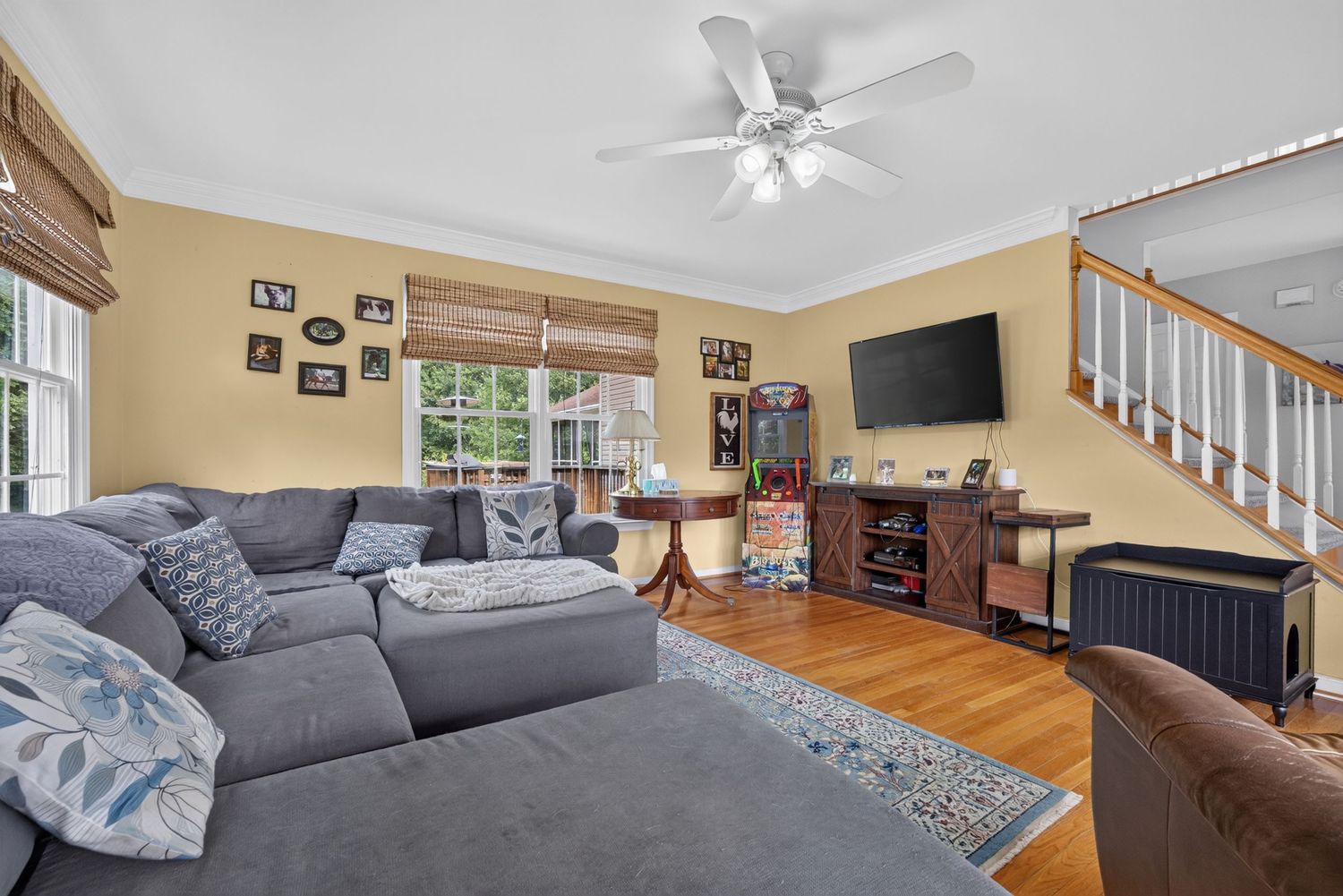 Cozy living room with gray sectional sofa, hardwood floors, ceiling fan, wall-mounted TV, and staircase in a bright home.