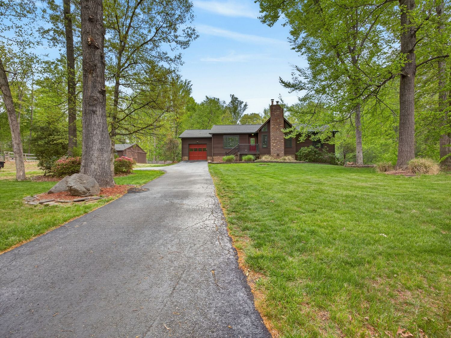 A charming ranch-style home with brick chimney, red garage, and long driveway surrounded by lush green lawn and tall trees.
