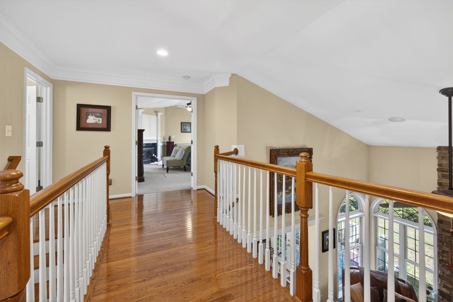 Bright upstairs hallway with hardwood floors, white spindle railing, crown molding, and views into a living room and outdoor deck.
