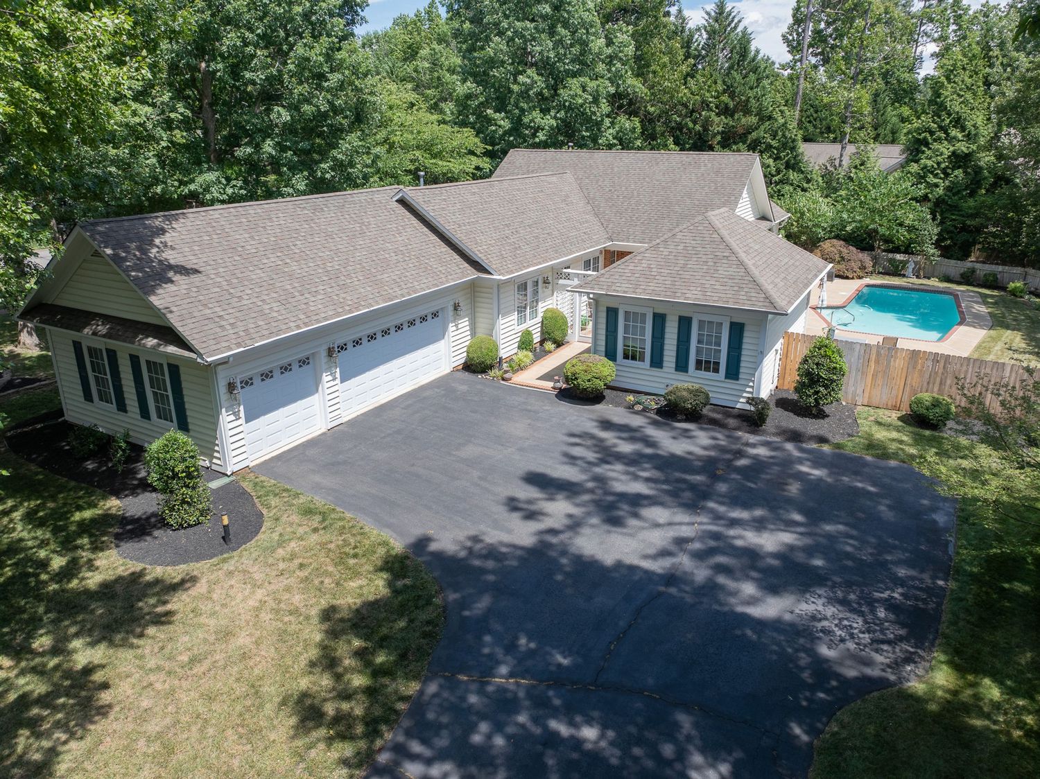 Aerial view of a spacious ranch-style home with a two-car garage, large driveway, lush trees, and a backyard pool.
