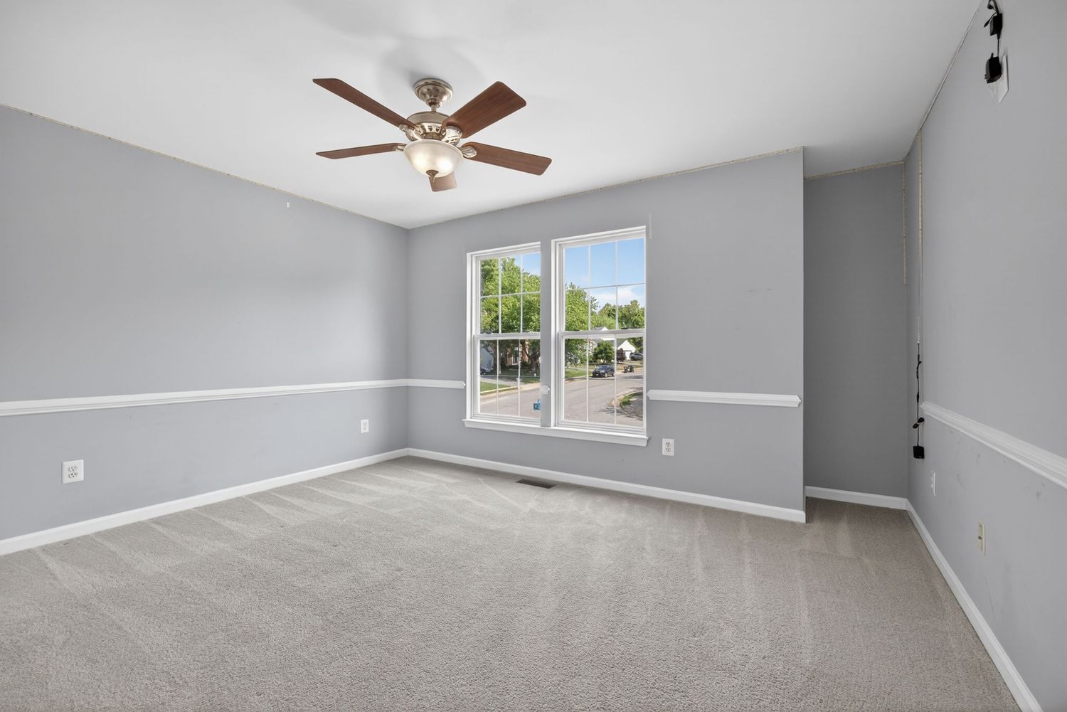 Empty room with gray walls, ceiling fan, and large window allowing natural light, featuring neutral carpet flooring.