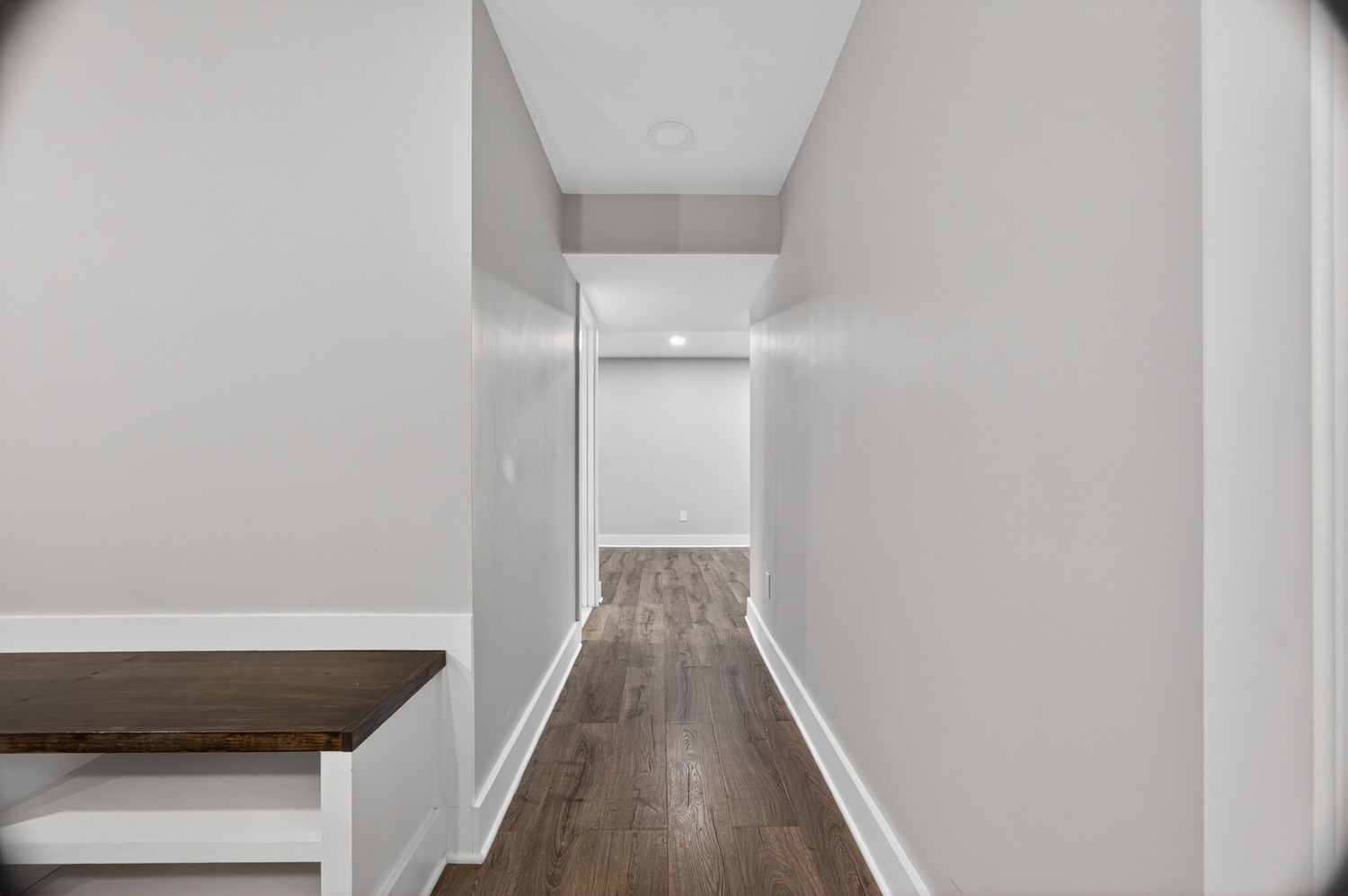 Narrow hallway with dark hardwood floors, white walls, and white baseboards leading to a bright open room.
