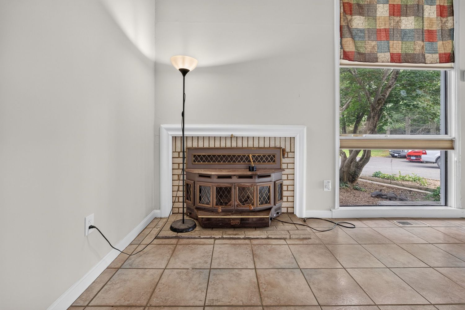 Tiled living room with ornate wood-burning stove fireplace, floor lamp, and large window overlooking lush greenery.