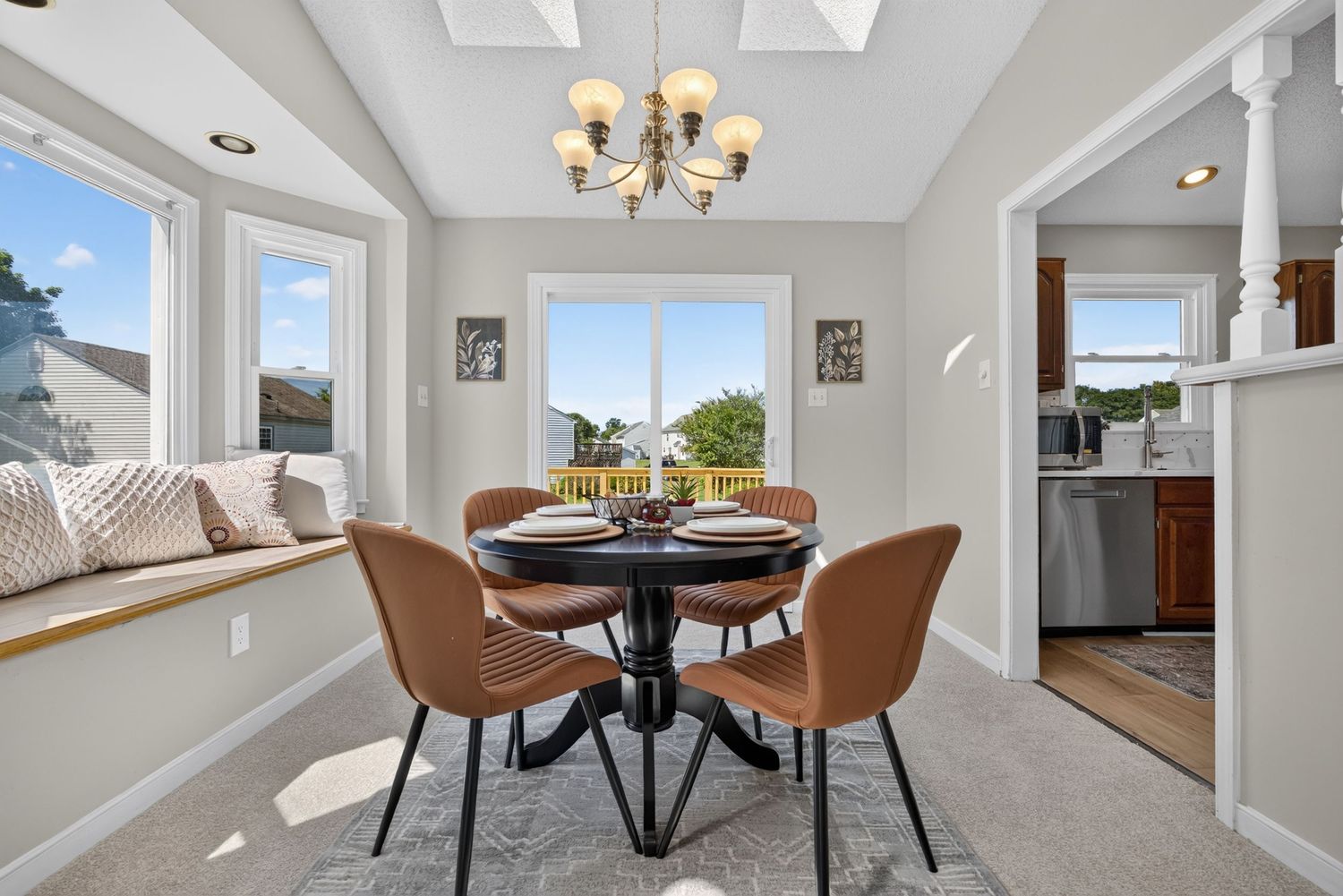 Bright dining room with round black table, brown leather chairs, chandelier, bay window seating, and view of outdoor scenery.