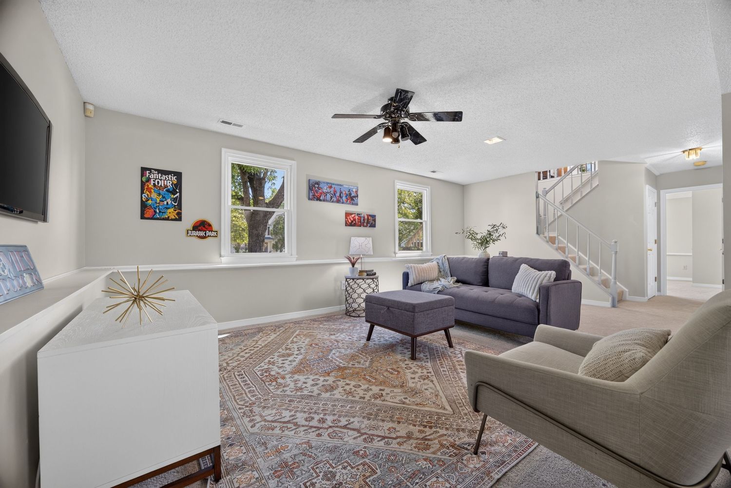 Bright living room with gray sectional sofa, patterned area rug, ceiling fan, and staircase in a modern home.