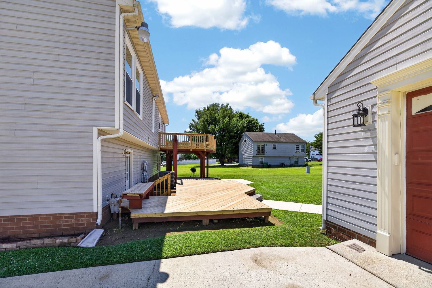 Suburban backyard with wooden deck, pergola, lush green lawn, and neighboring homes under a bright blue sky.