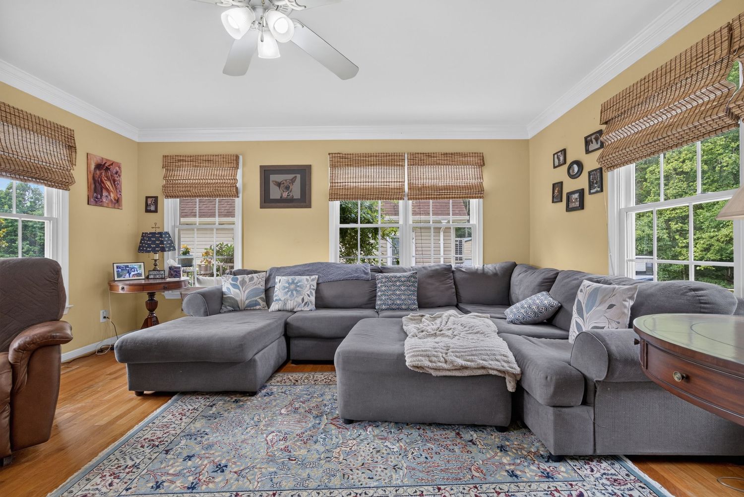 Spacious living room with gray sectional sofa, hardwood floors, blue area rug, bamboo shades, and ceiling fan.