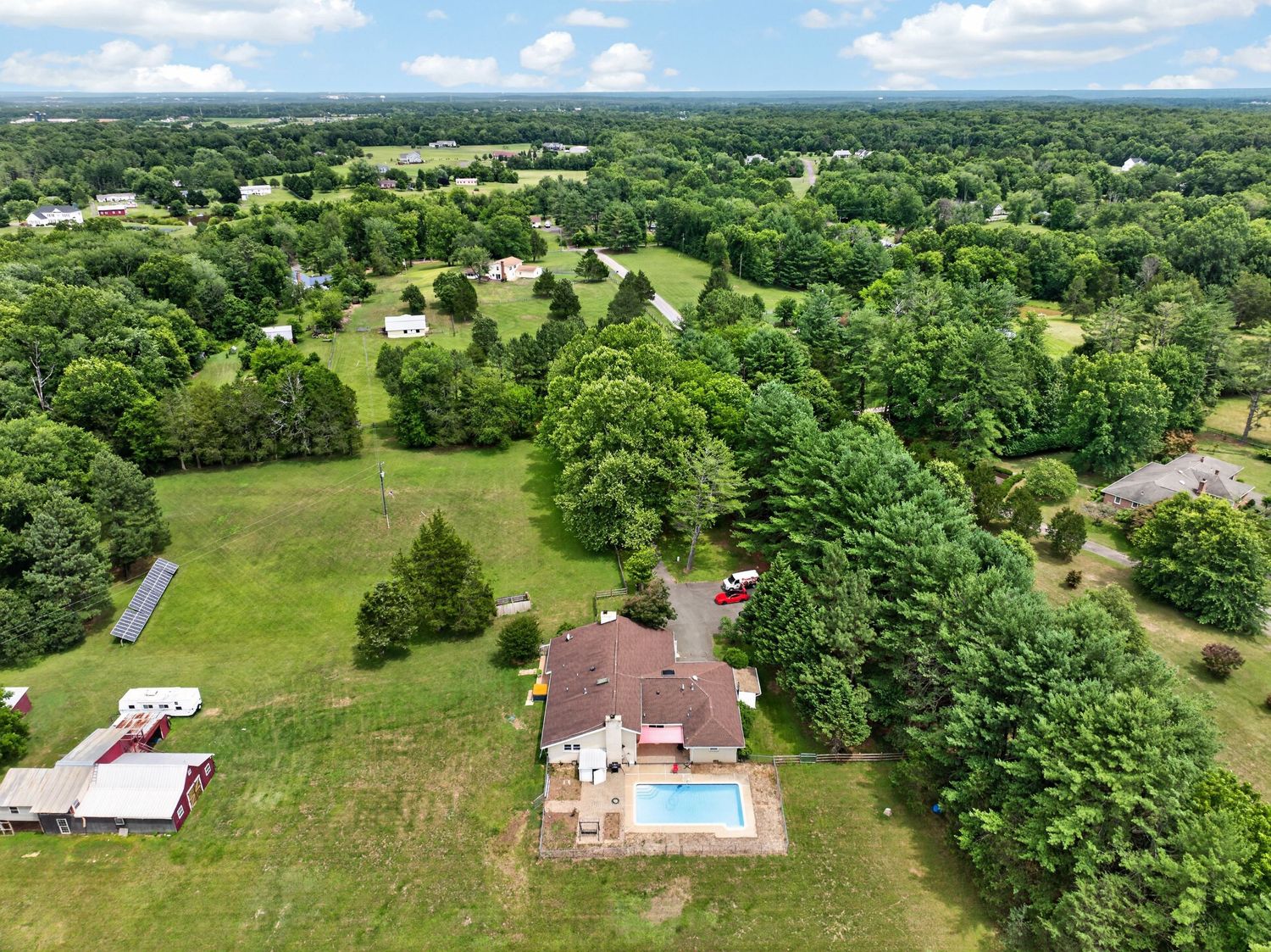 Aerial view of a suburban home with a swimming pool surrounded by lush green trees and open fields.