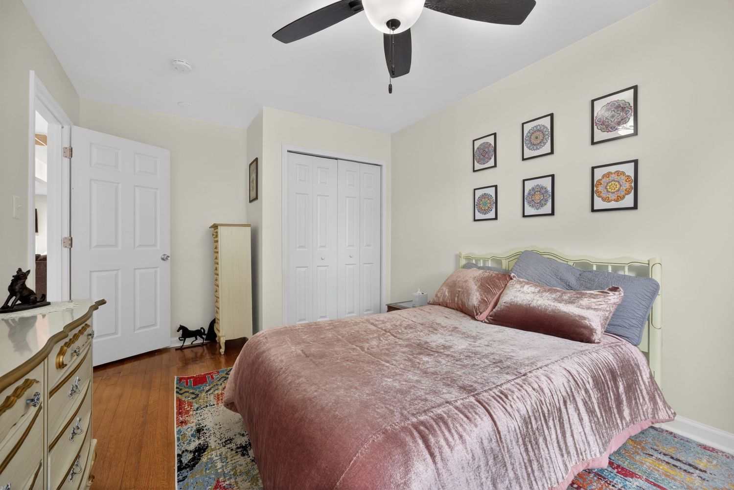 Cozy bedroom with pink velvet bed, hardwood floors, ceiling fan, and colorful area rug.