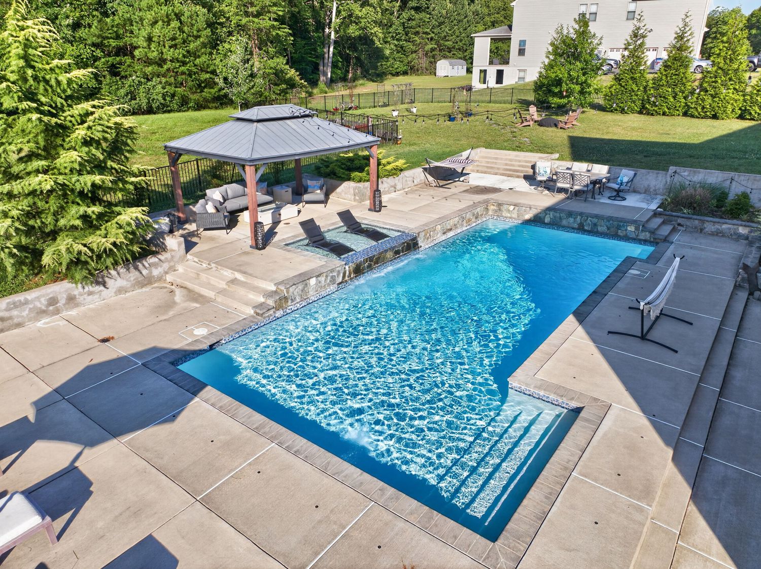 Aerial view of a rectangular swimming pool with a gazebo, lounge chairs, and lush green landscaping in a backyard.