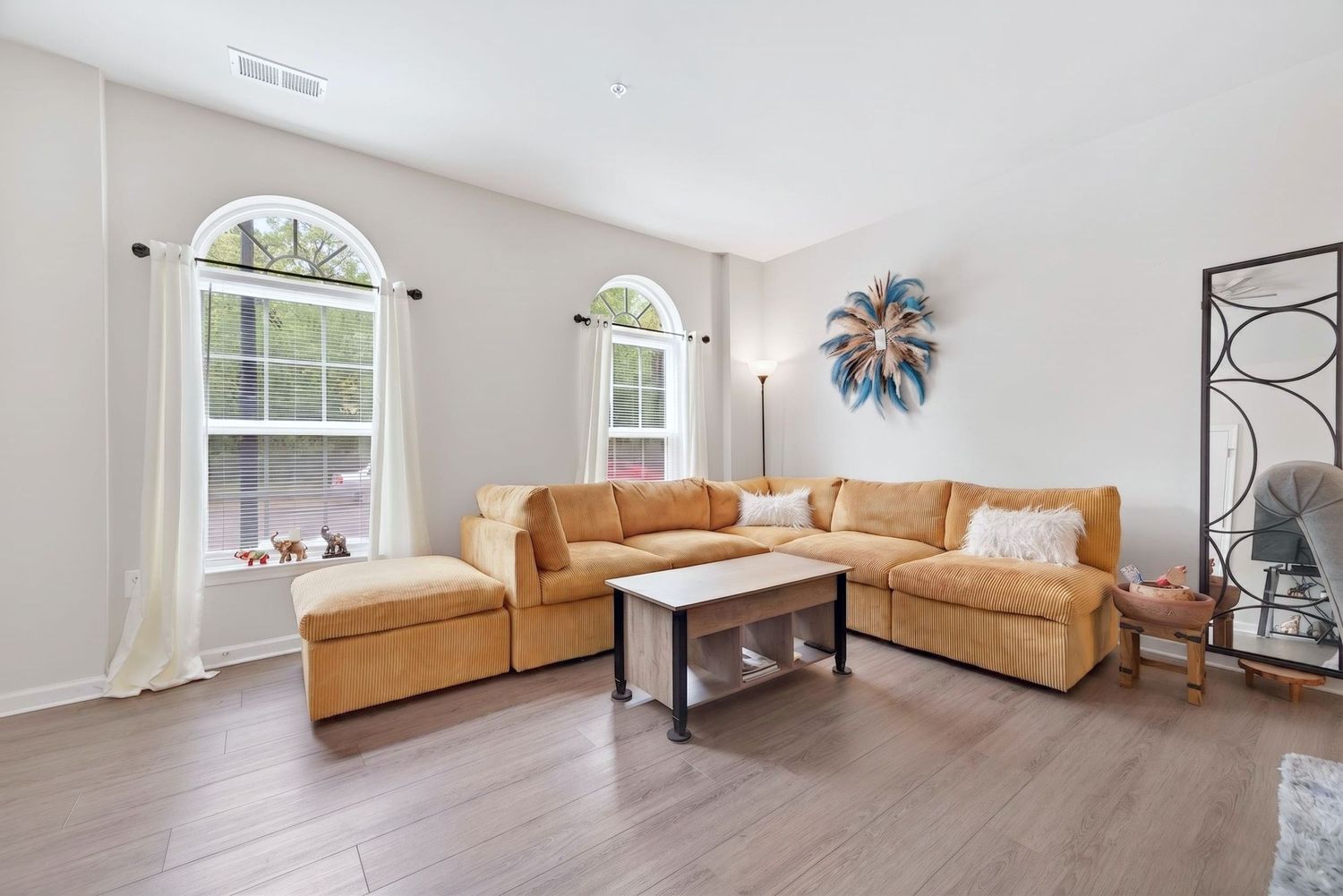 Bright living room with golden sectional sofa, white coffee table, and arched windows on light hardwood floors.
