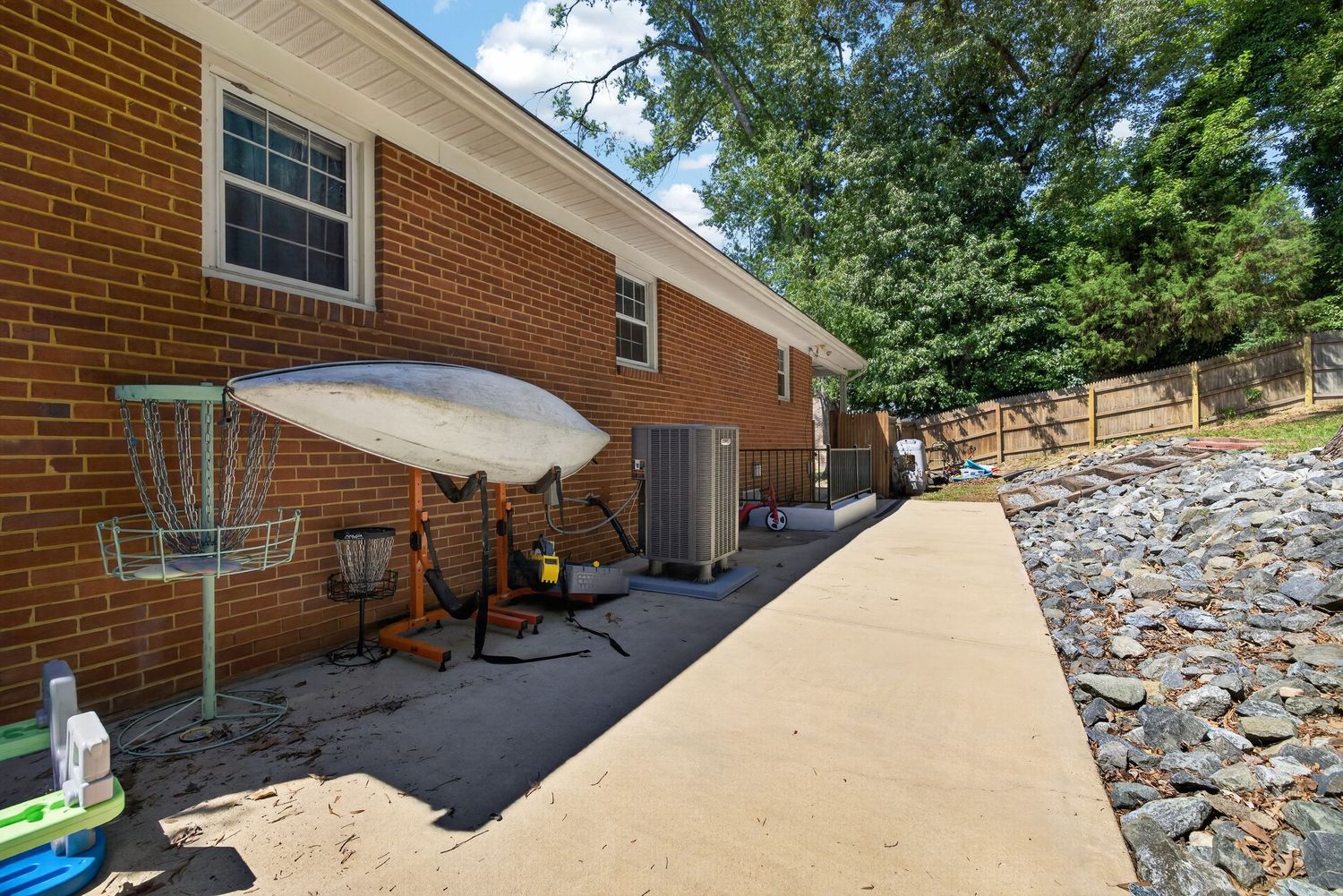 Brick ranch home exterior with concrete patio, disc golf basket, AC units, and gravel landscaping border surrounded by trees.
