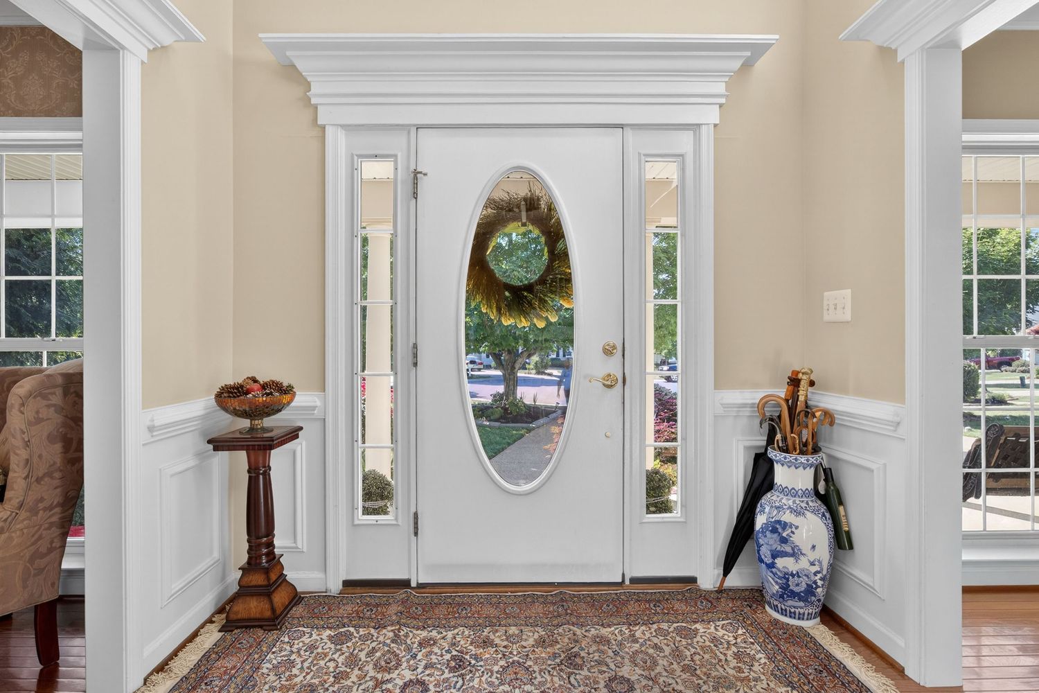 Elegant home entryway with white front door, oval glass panel, wreath, wainscoting, hardwood floors, and decorative vase.