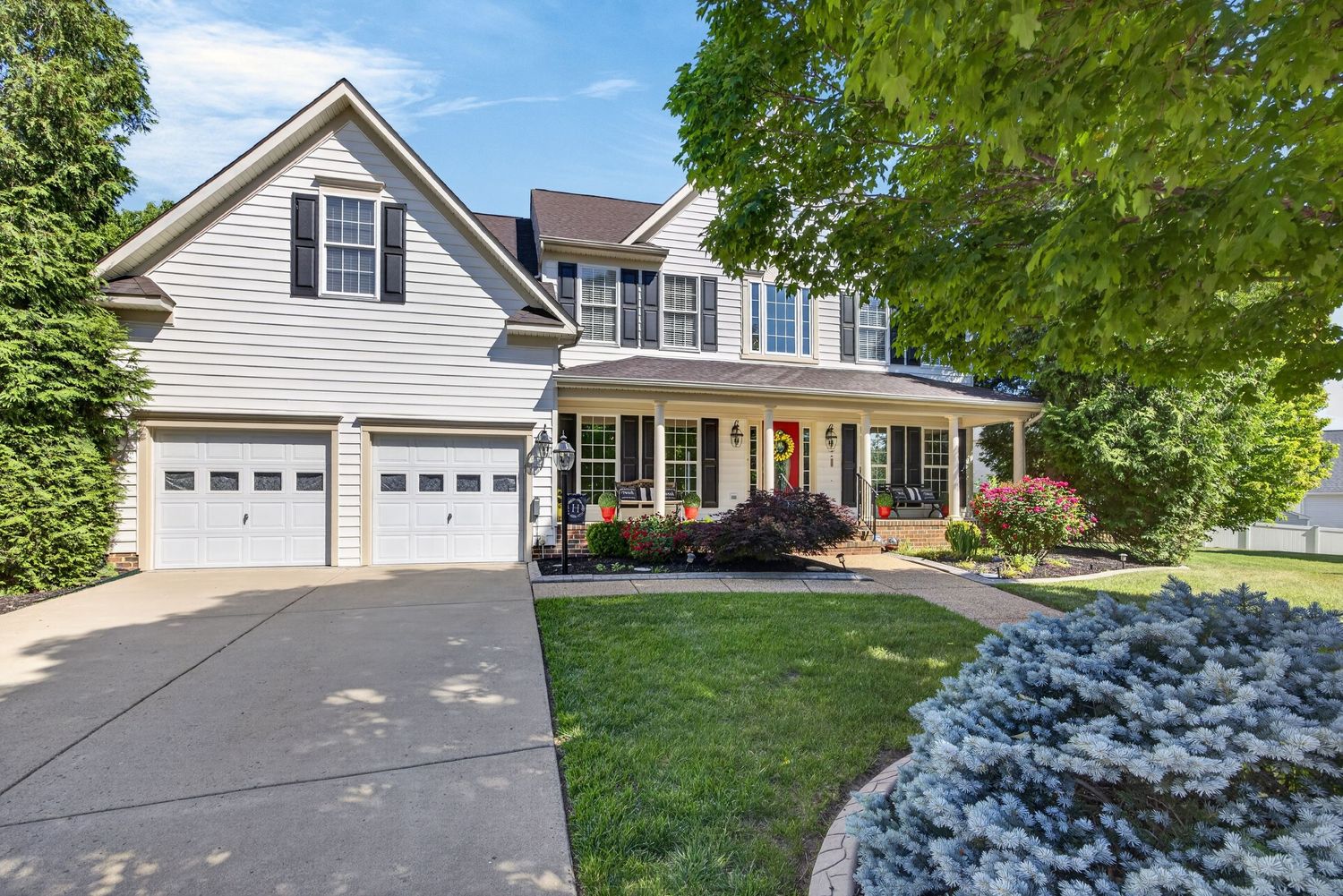Two-story colonial home with covered porch, two-car garage, manicured lawn, and mature trees on a sunny day.