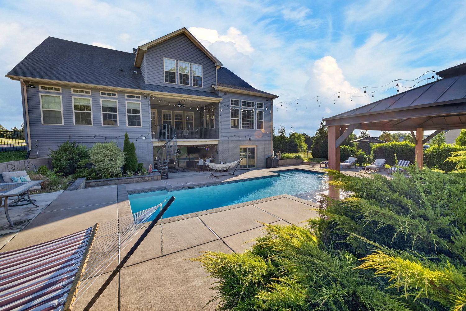 Large gray two-story home with rectangular pool, gazebo, and lush landscaping on a sunny day.