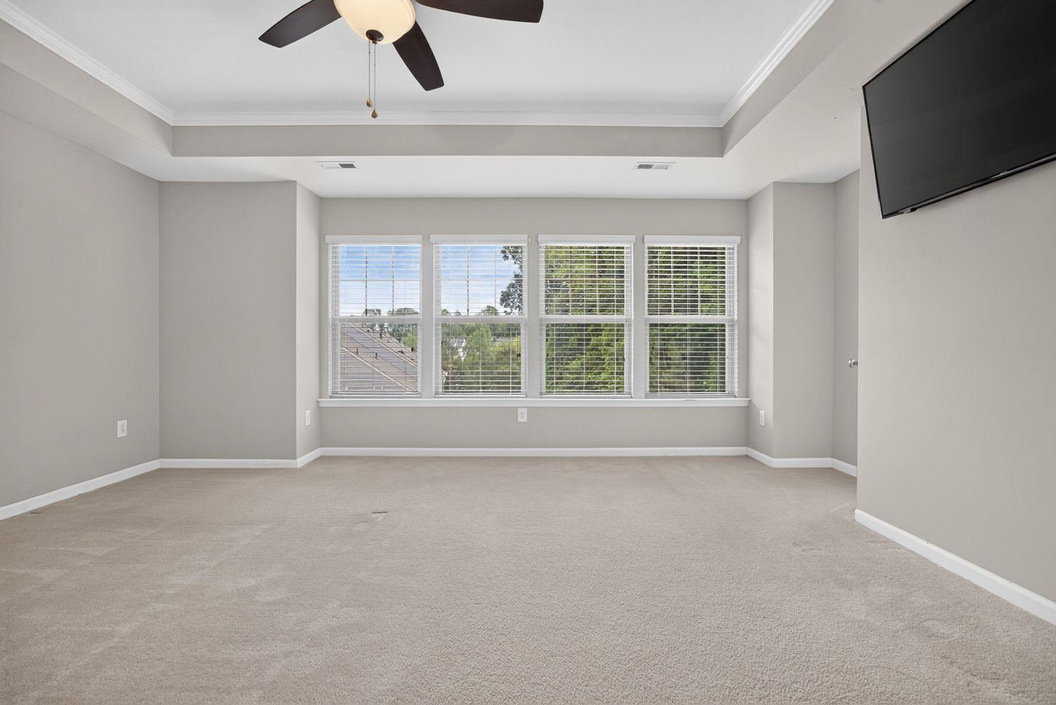 Bright empty bedroom with tray ceiling, ceiling fan, large windows, wall-mounted TV, and neutral carpet flooring.
