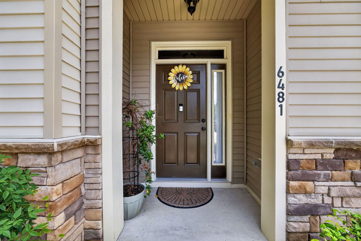 Welcoming front door entry with stone accents, sunflower wreath, and house number 6481 on a beige home.