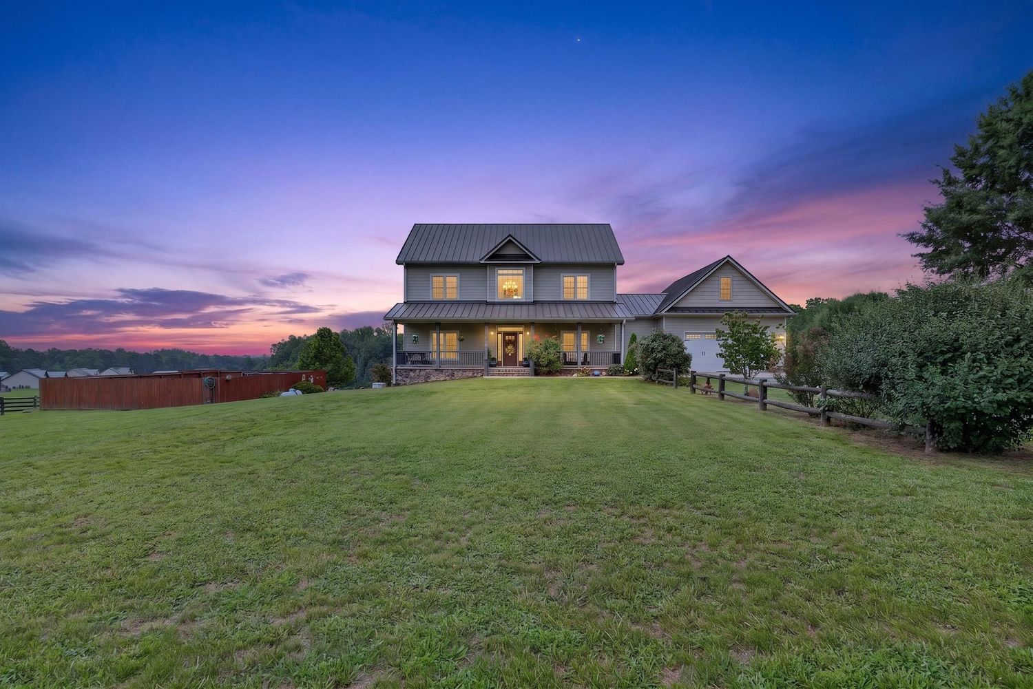 Two-story farmhouse with wrap-around porch sits on lush green lawn at twilight with colorful purple and orange sunset sky.
