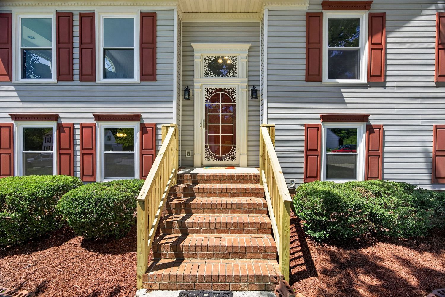Brick steps lead to a white front door with decorative glass, flanked by red shutters and lush green shrubs.