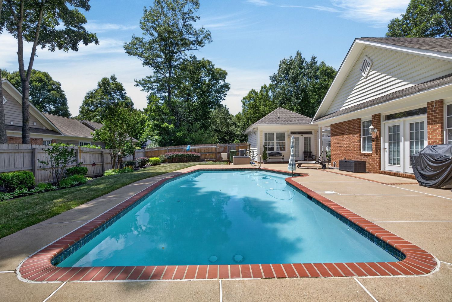 Rectangular backyard swimming pool with brick border, concrete patio, and lush trees surrounding a brick home.