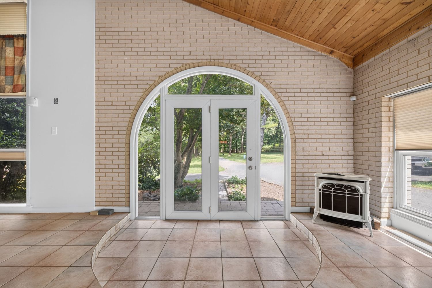 Sunlit sunroom with arched white French doors, brick walls, tile flooring, and vaulted wood ceiling leading to a lush backyard.