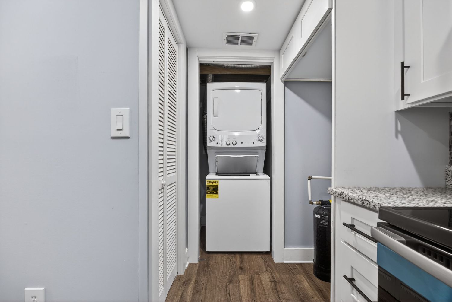 Hallway with stacked washer and dryer unit, white cabinetry, dark hardwood floors, and granite countertops in adjacent kitchen.