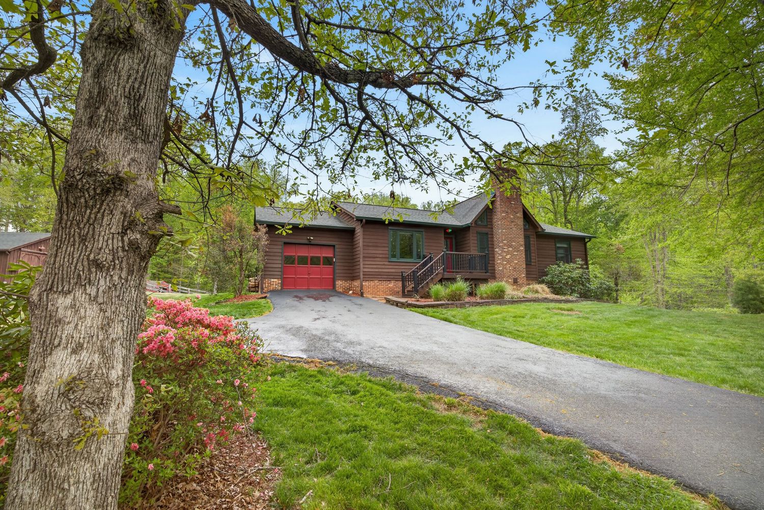 Charming wood-sided ranch home with red garage door, lush green lawn, and blooming azaleas along a paved driveway.