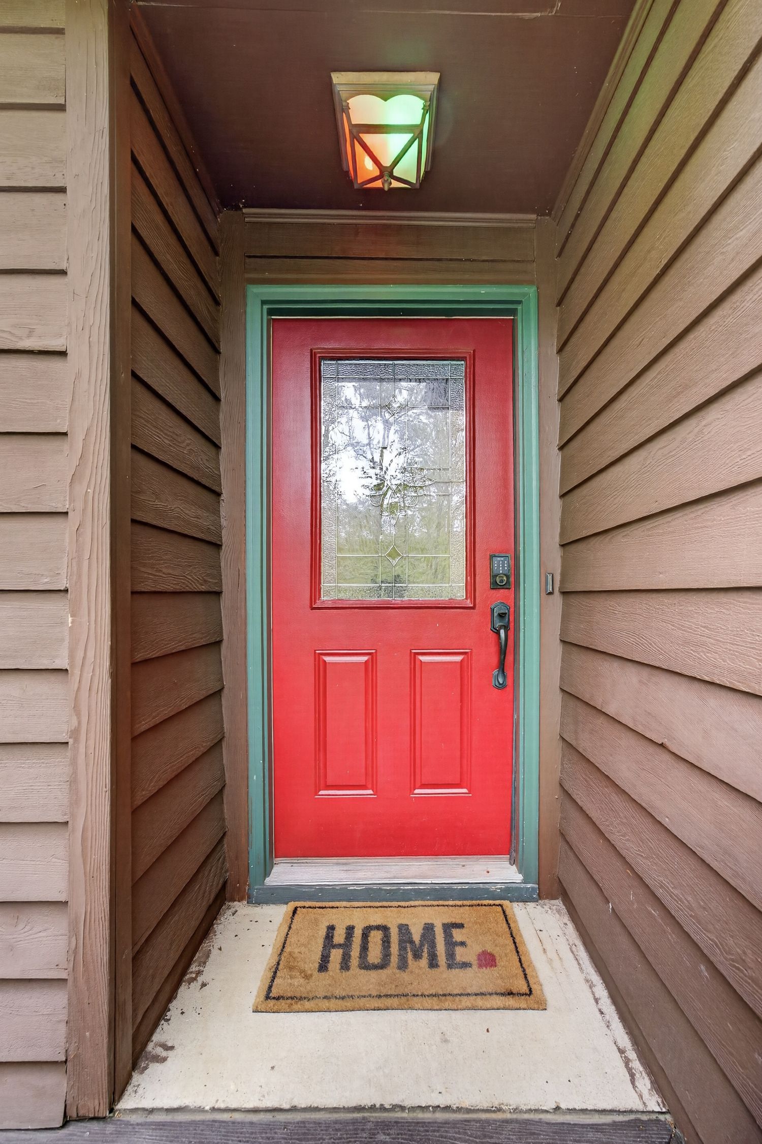 Red front door with green trim, glass panel, and a HOME doormat on a covered porch with wood siding.