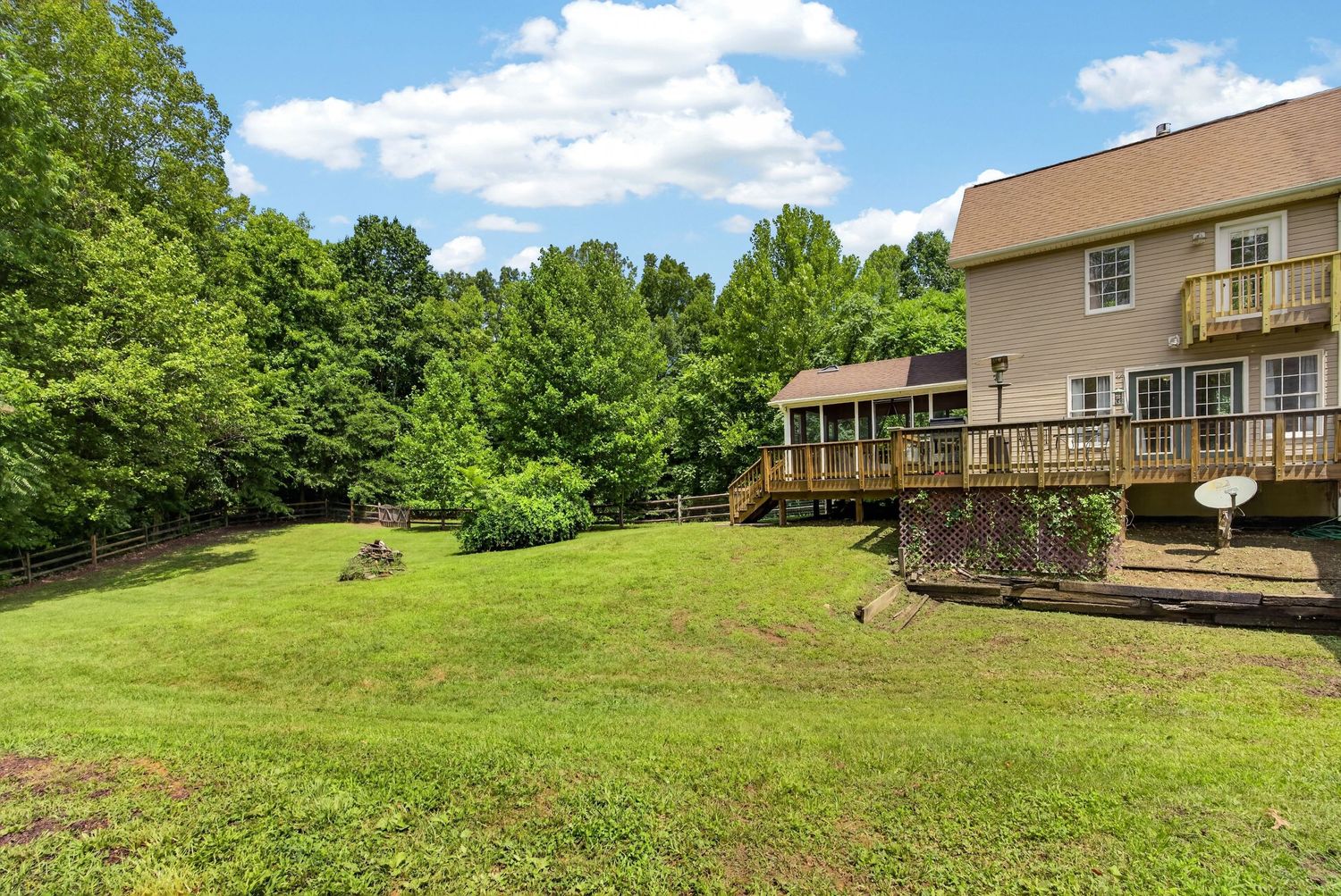Spacious backyard with lush green lawn, large wooden deck, and surrounding mature trees on a sunny day.
