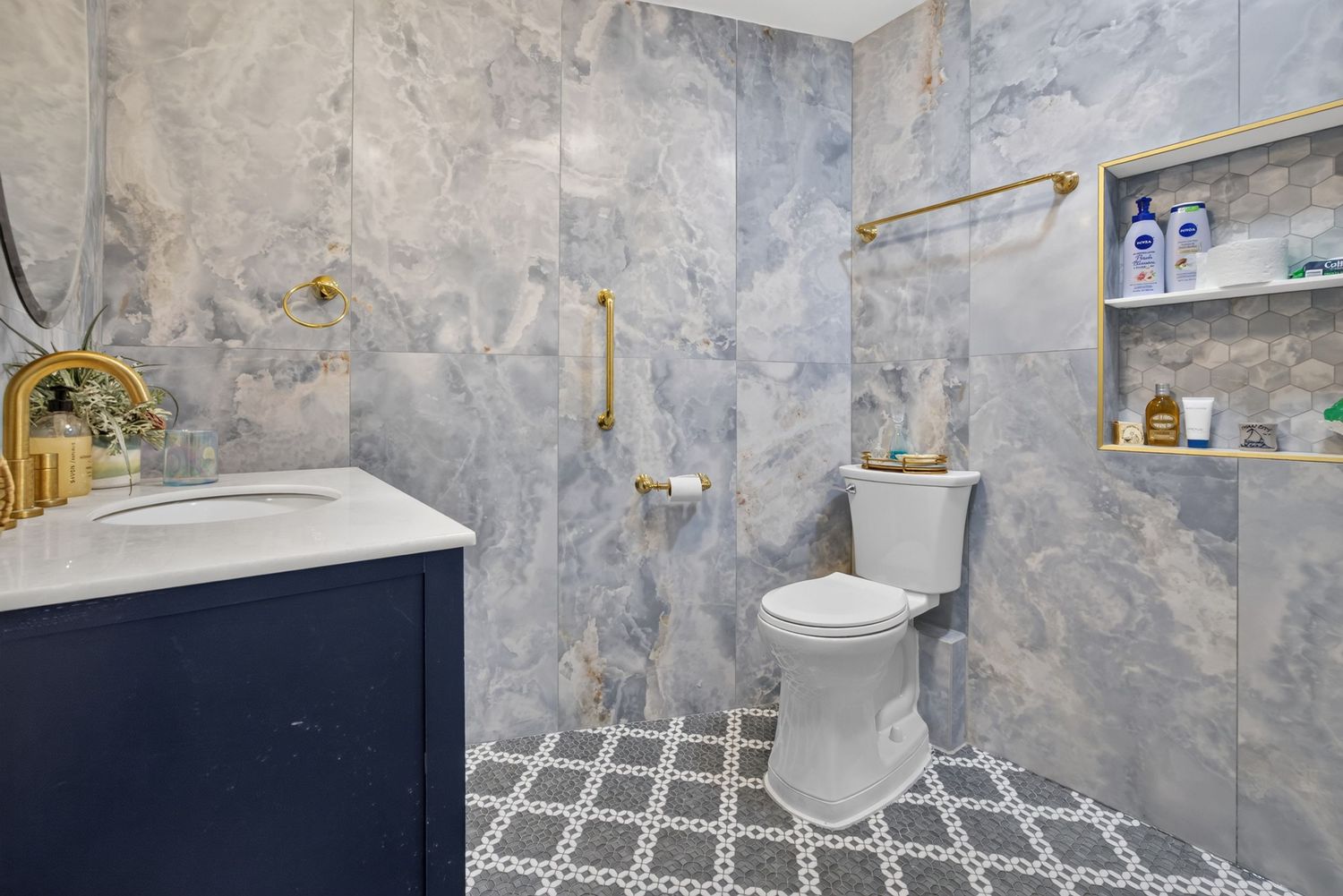 Modern bathroom with gray marble walls, gold fixtures, patterned tile floor, navy vanity, and built-in shelving niche.