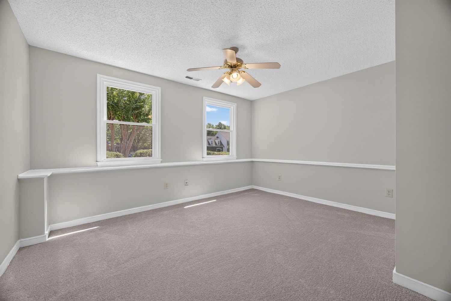 Empty bedroom with gray carpet, ceiling fan, and two windows with natural light in a clean, neutral-toned room.