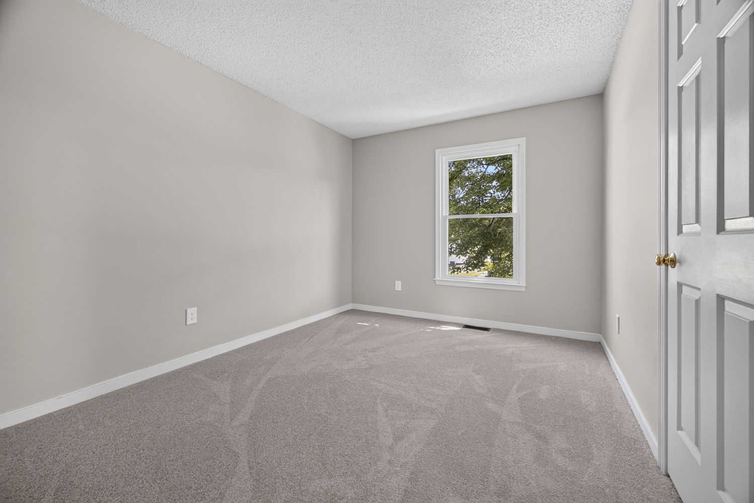 Empty bedroom with gray carpet, white walls, a window with natural light, and a white door in a clean, bright space.