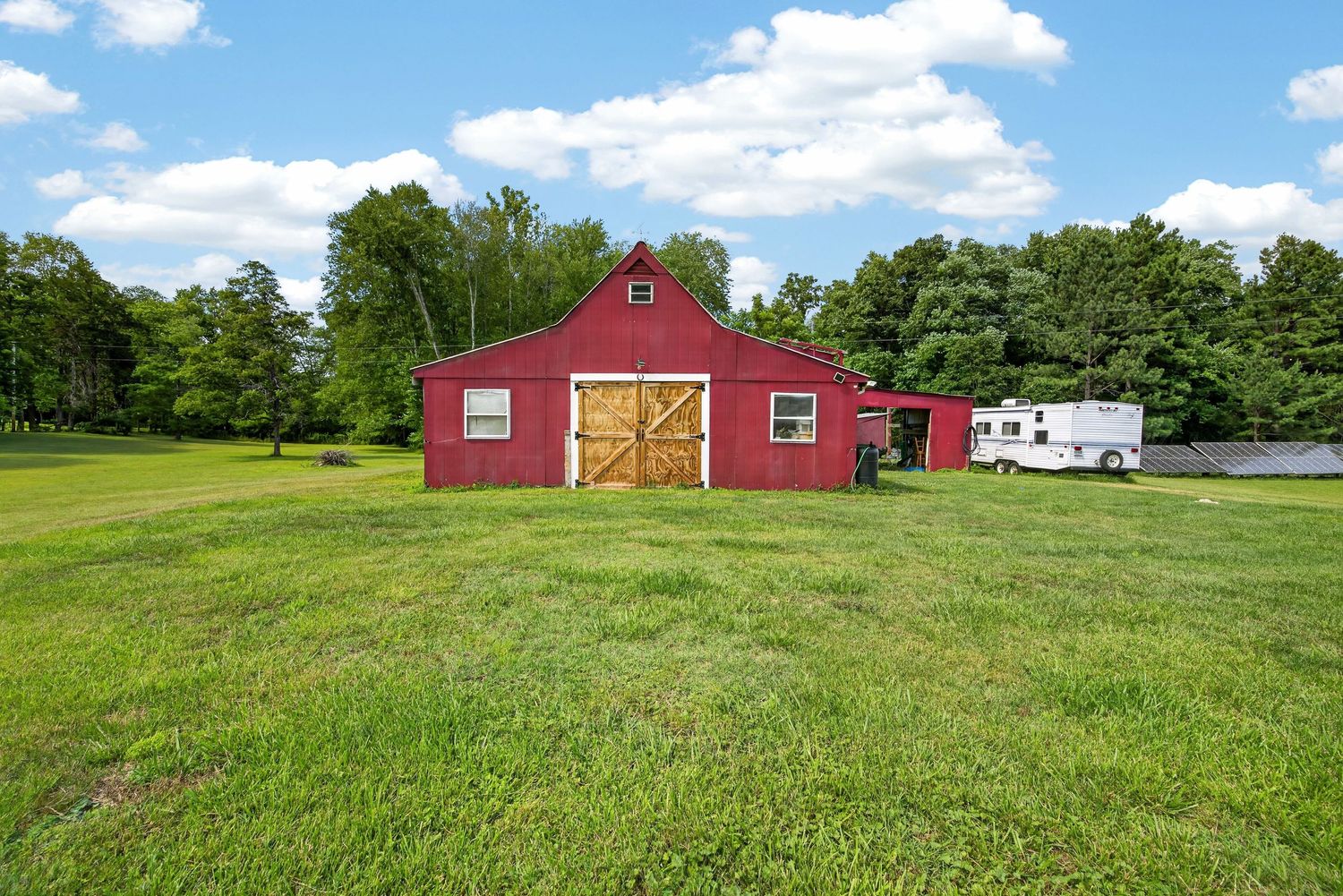 Red barn with wooden doors on a lush green rural property, surrounded by trees with a trailer parked nearby.