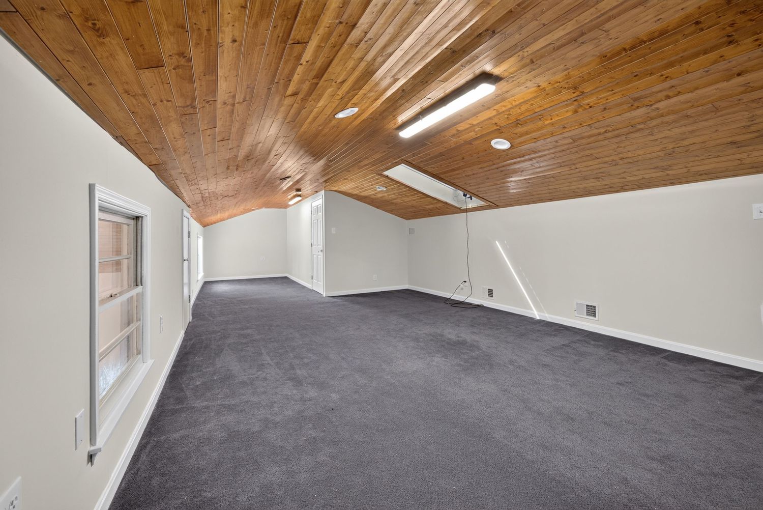 Spacious room with vaulted wood ceiling, gray carpet, white walls, and fluorescent lighting in an attic-style space.