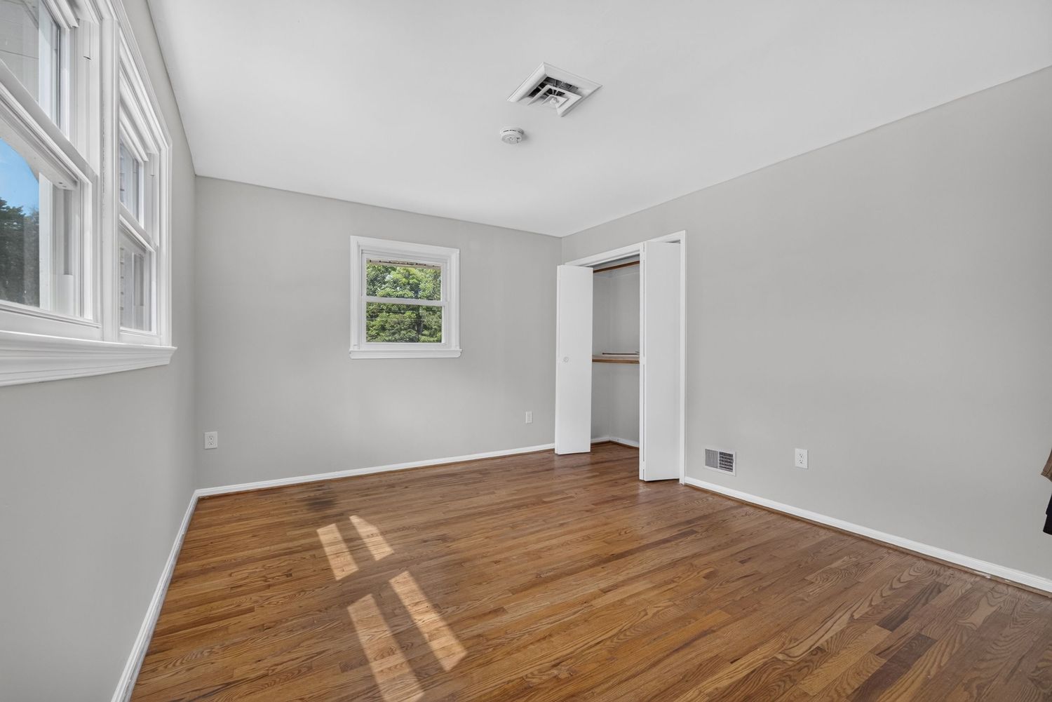 Bright empty bedroom with hardwood floors, gray walls, white trim, closet, and two windows allowing natural light.
