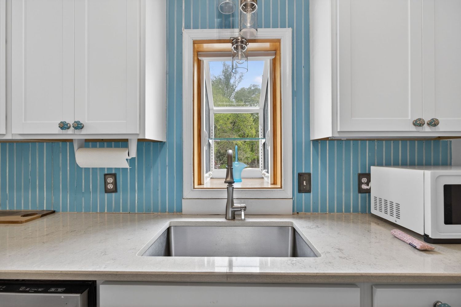 Modern kitchen sink with stainless steel basin, blue beadboard backsplash, white cabinets, and a window overlooking greenery.