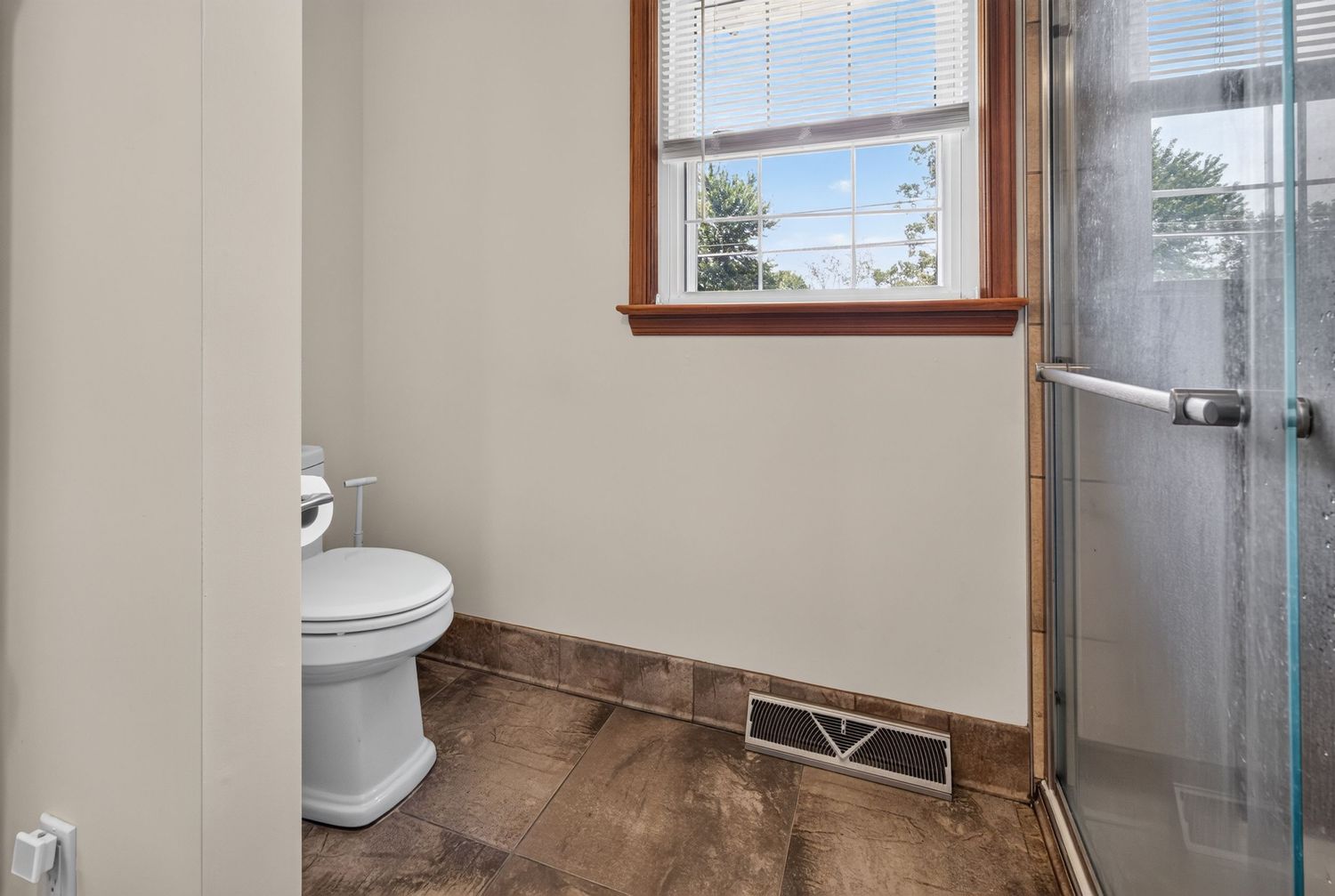 Bathroom with white toilet, glass shower door, dark tile floors, and a window providing natural light.