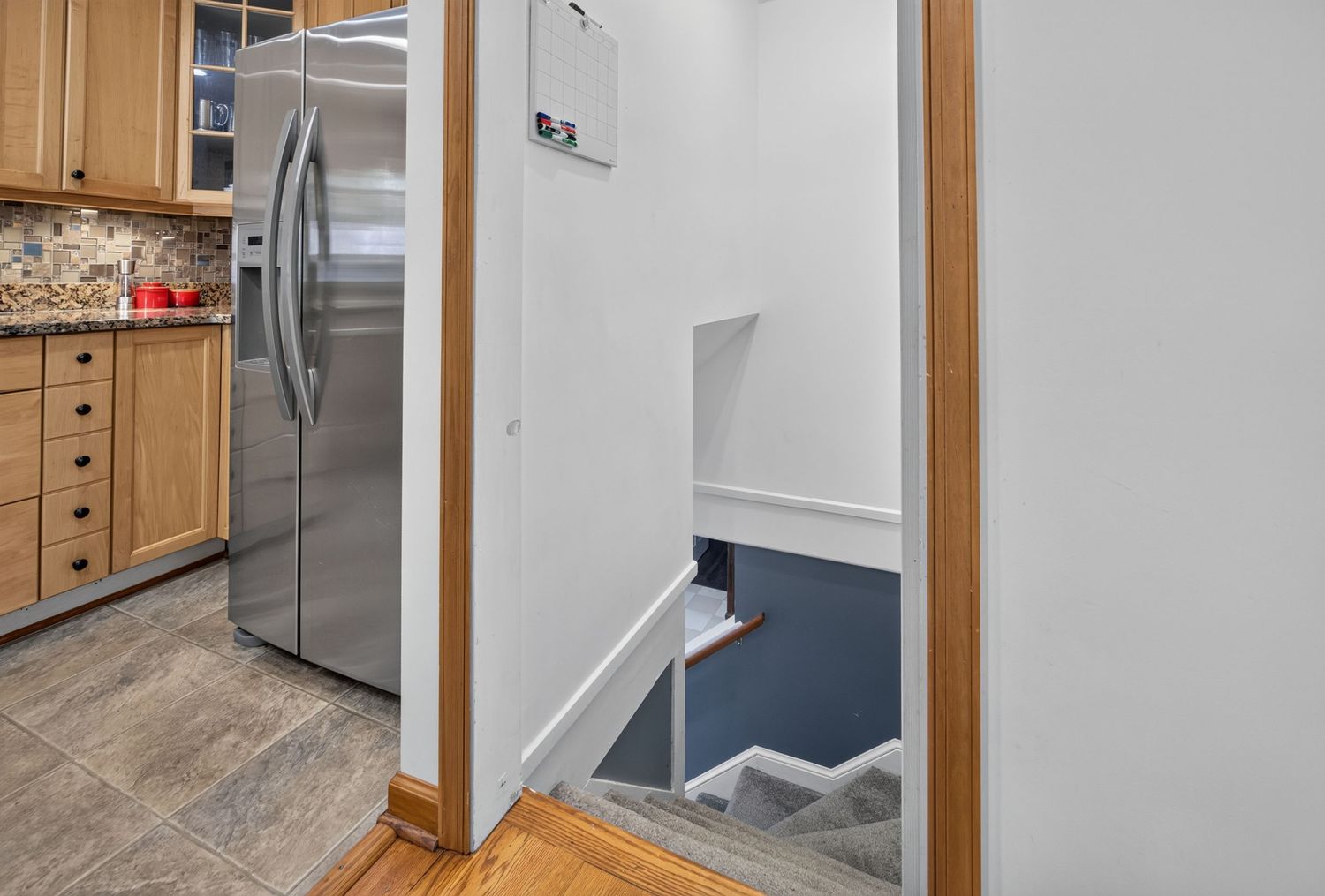 Staircase leading to lower level beside a modern kitchen with stainless steel refrigerator and wooden cabinetry.