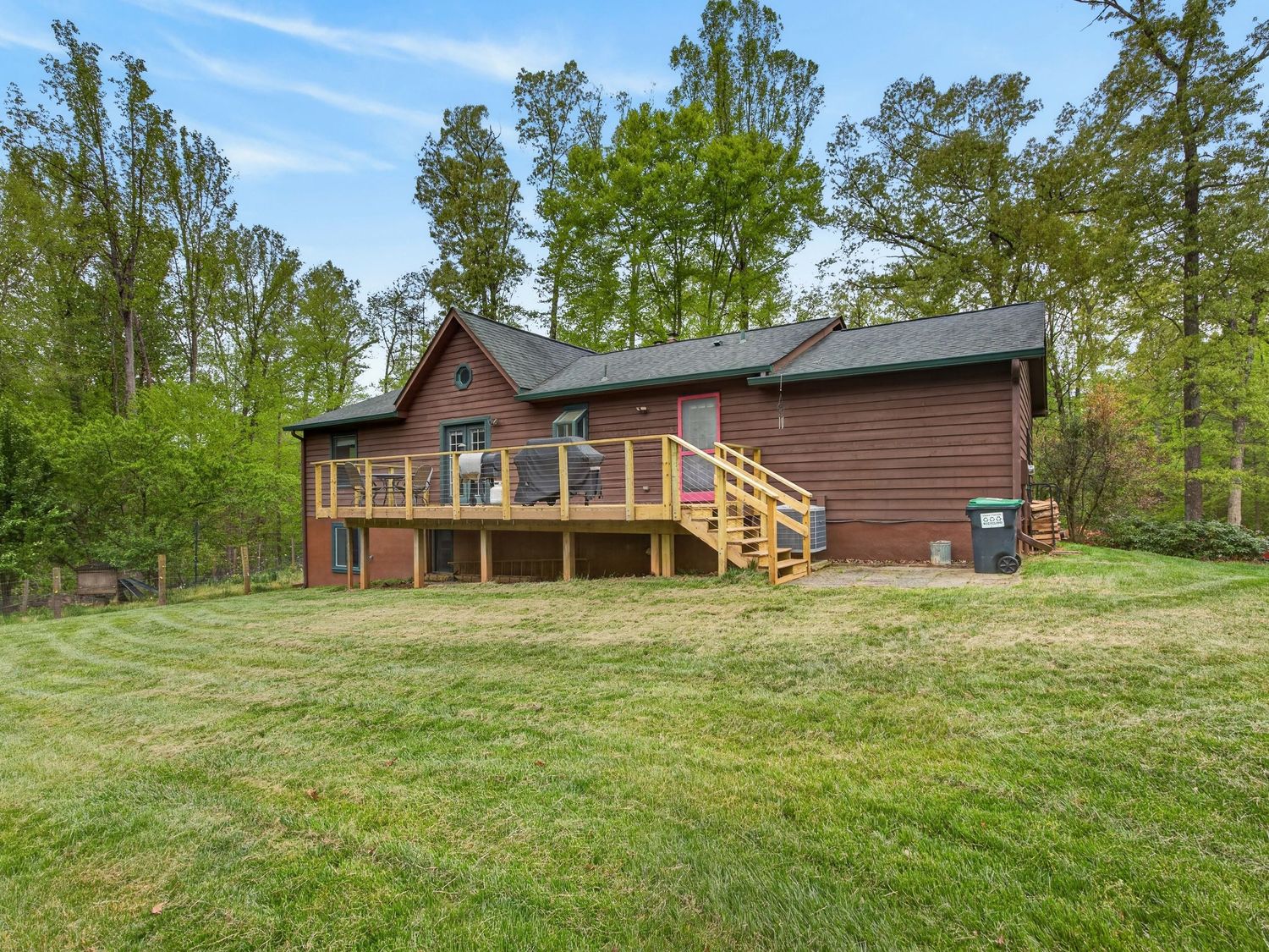 Rustic brown cabin with large wooden deck surrounded by lush green lawn and tall trees in a serene wooded setting.