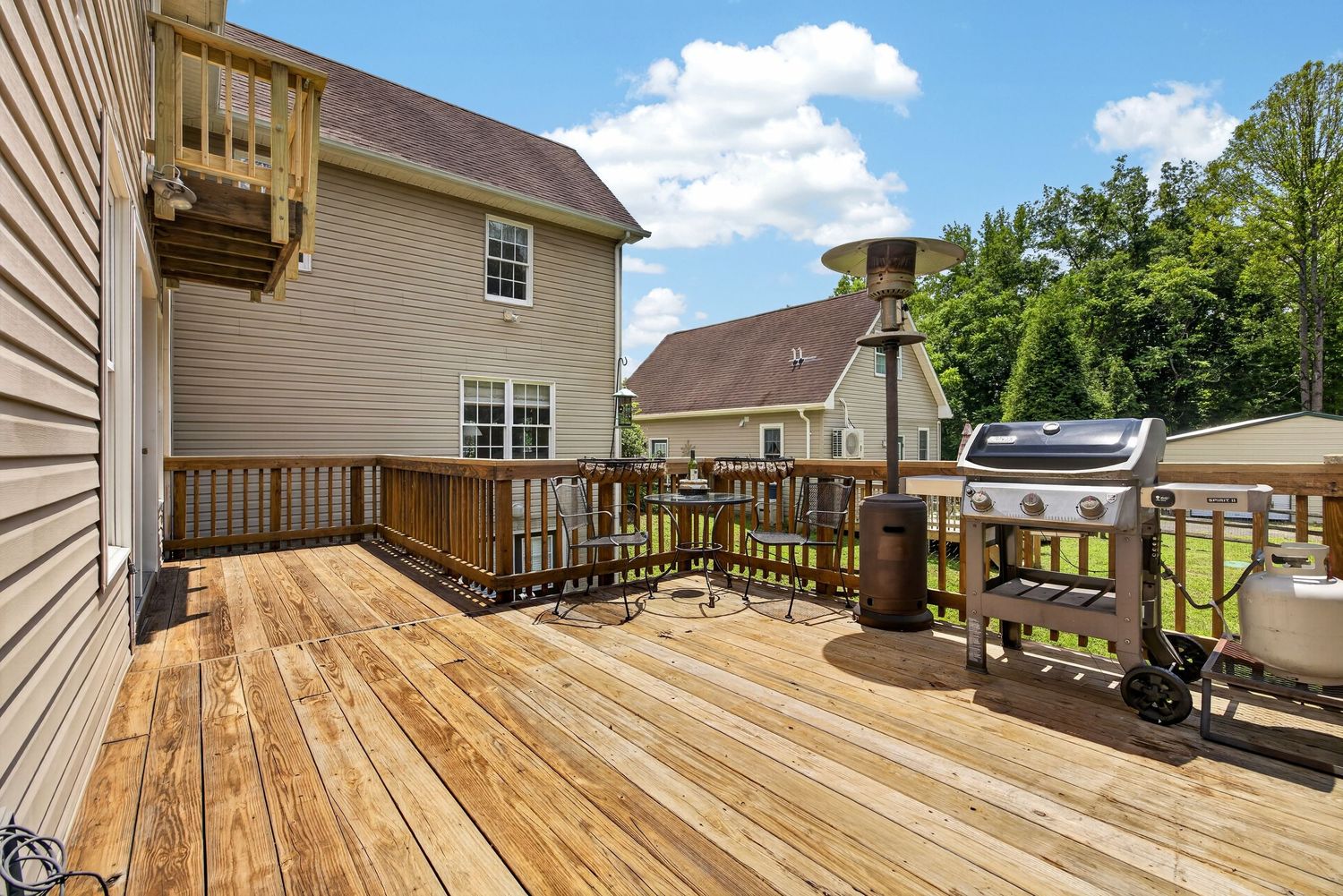 Spacious wooden deck with gas grill, multi-level design, and lush green trees in background on sunny day.