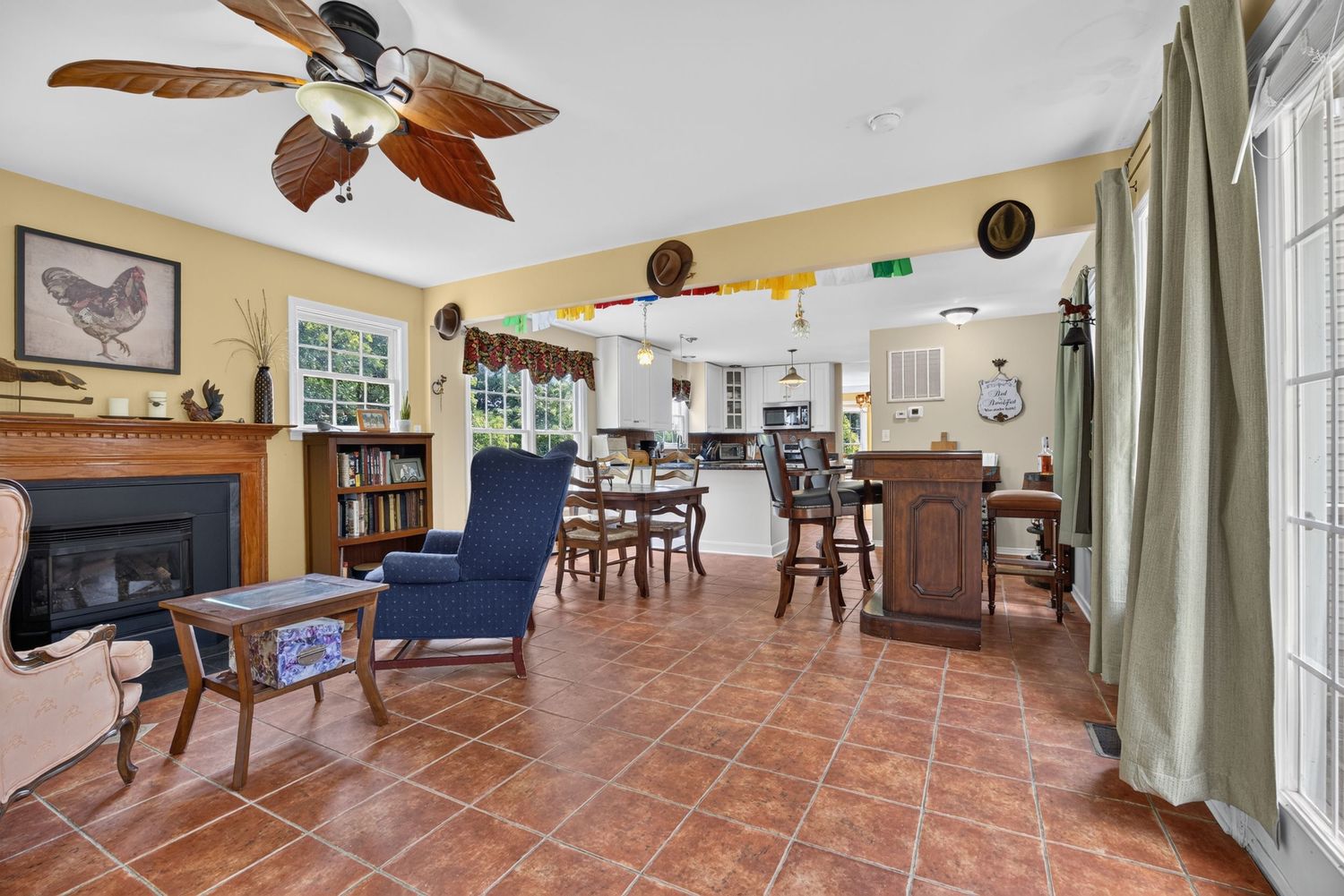 Spacious living room with terracotta tile floors, ceiling fan, fireplace, blue armchair, and open view to dining area and kitchen.