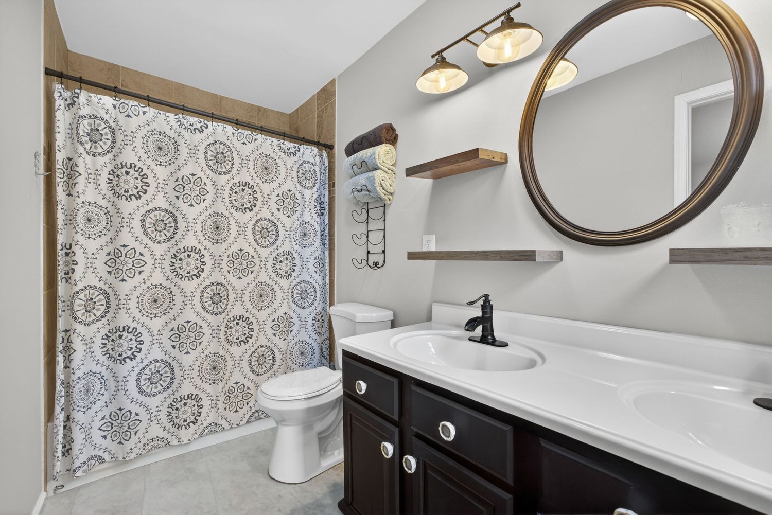Modern bathroom with dark wood vanity, round mirror, patterned shower curtain, and white countertop with black faucet.