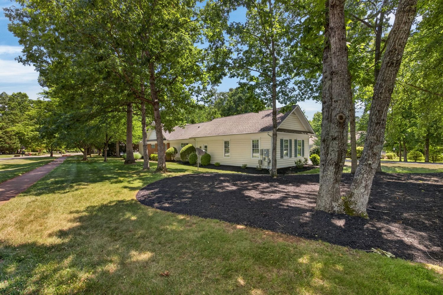 Yellow cottage home surrounded by tall mature trees with a dark mulched driveway and lush green lawn on a sunny day.